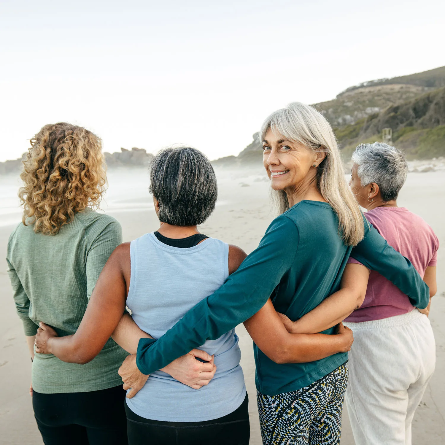 Vier ältere Damen, die einen kleinen Halbkreis bilden, sich umarmen und eine Dame dreht sich um und lächelt in die Kamera.